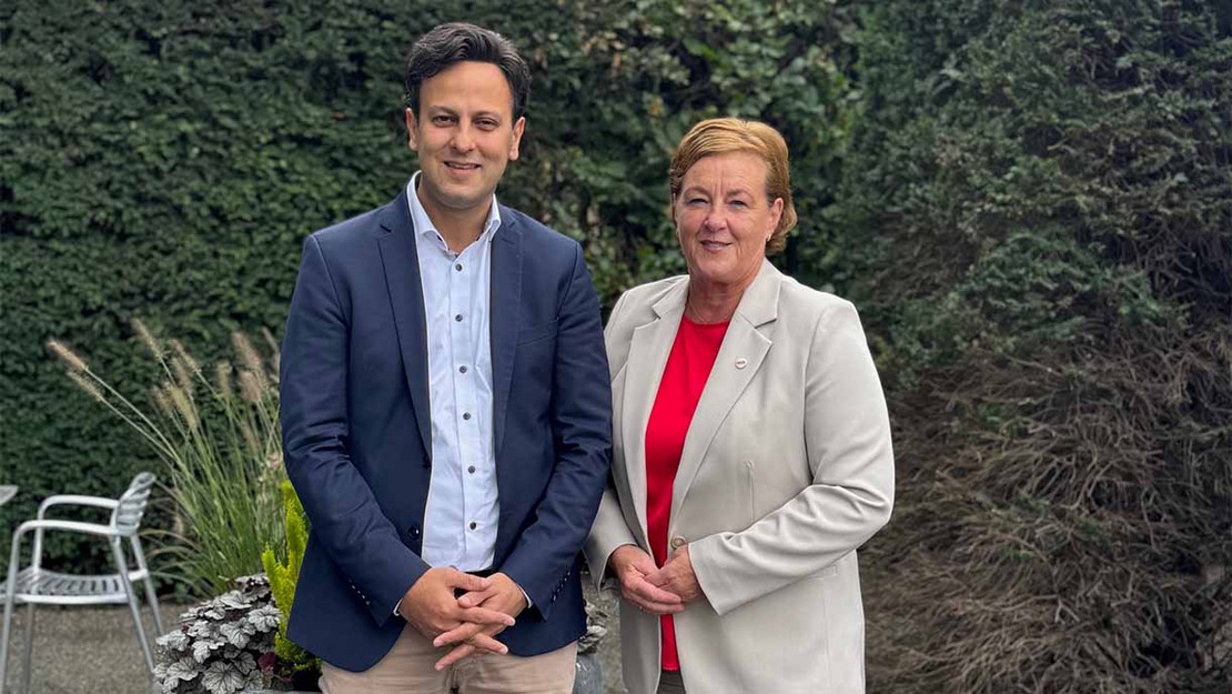 Zwei Personen stehen nebeneinander, im Hintergrund eine grüne Hecke. 