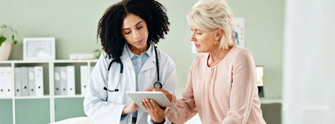 Eine Ärztin hält ein Tablet in der Hand und zeigt einer Patientin etwas darauf. 