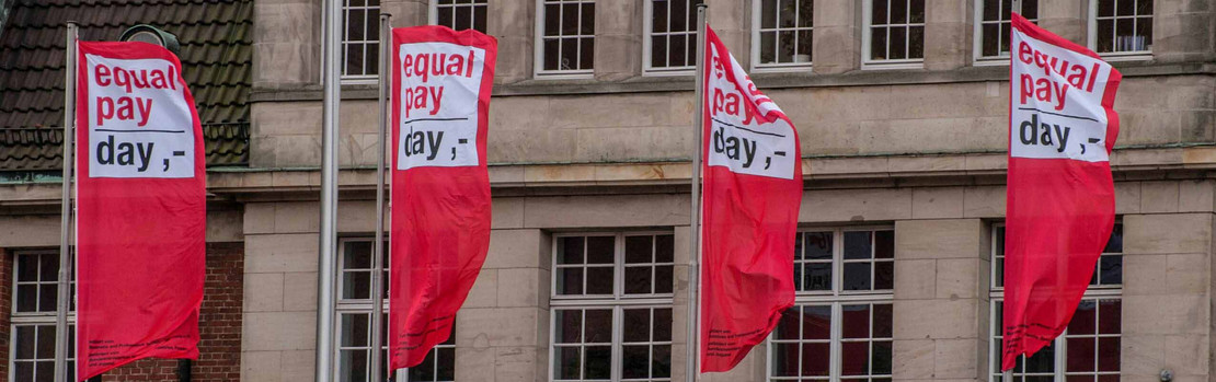Zwei Flaggen mit dem Equal-Pay-Day-Logo wehen vor einem Gebäude. 