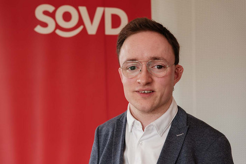 Porträtfoto: Junger Mann vor einer roten Wand mit SoVD-Logo. 