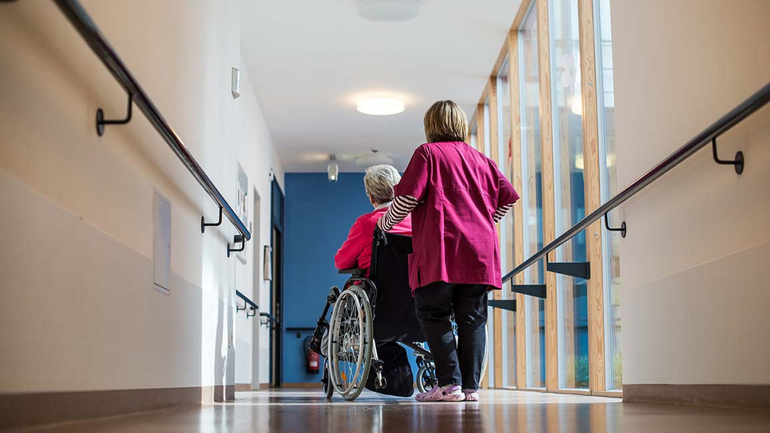 Eine Frau schiebt eine ältere Frau in einem Rollstuhl durch den Gang eines Pflegeheims. 