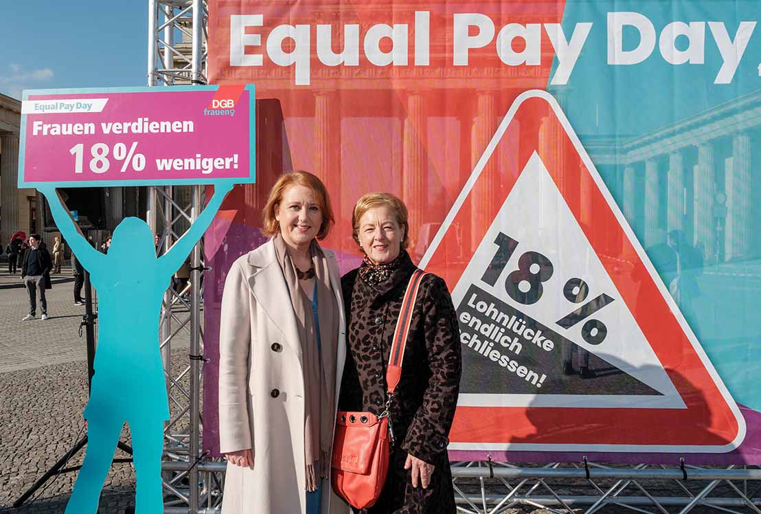 Zwei Frauen vor einem Plakat mit der Aufschrift "Equal Pay Day" und "Frauen verdienen 18 Prozent weniger". 