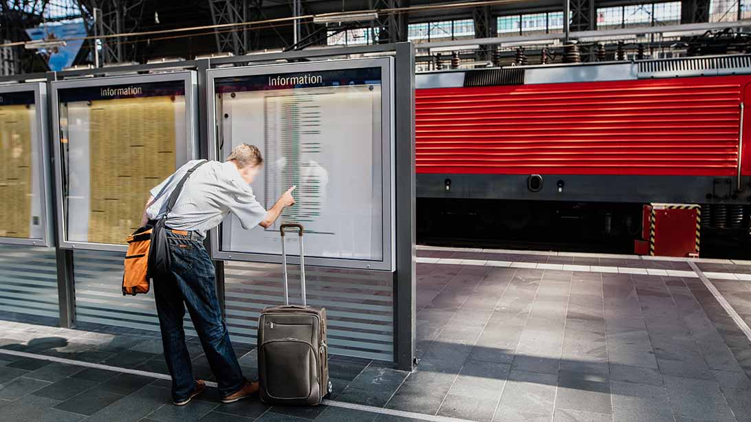 Mann steht am Bahnhof und schaut auf den Informationstafeln nach seinem Zug. 