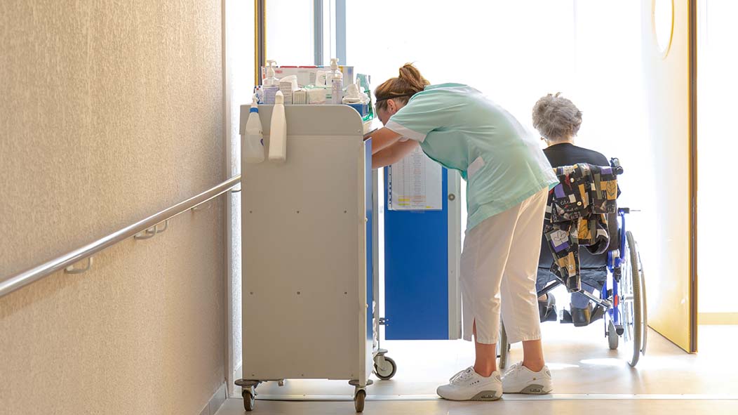 Eine Pflegekraft beugt sich über einen Medikamentschrank, im Hintergrund sitzt eine ältere Frau im Rollstuhl. 